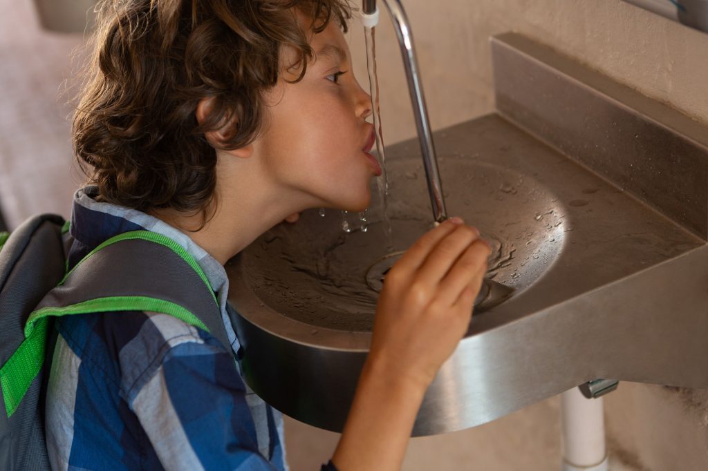 Side view of a Caucasian schoolboy drinking water from tap in the corridor at school